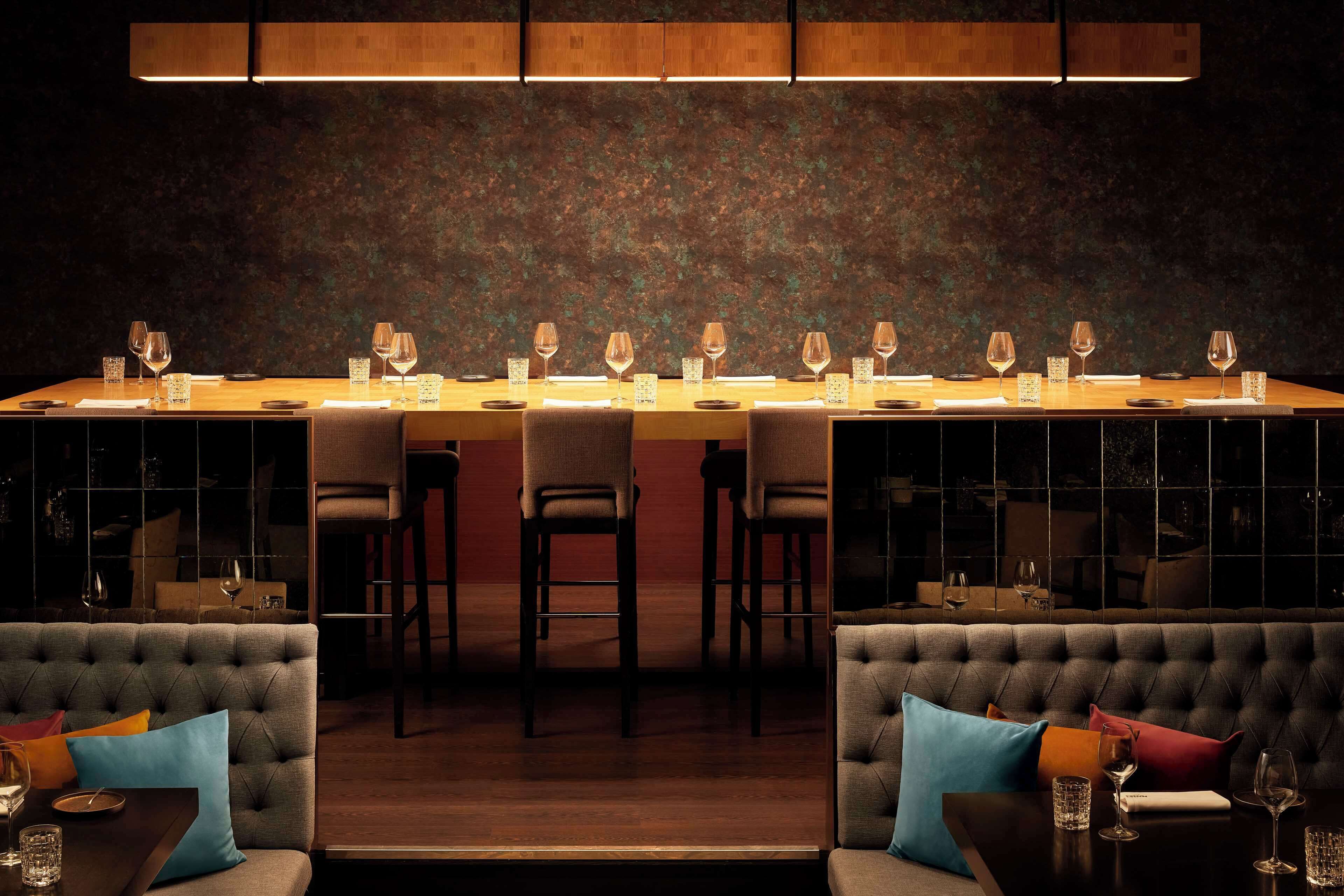 A restaurant with a long wooden bar, cushioned seating, and a textured wall.