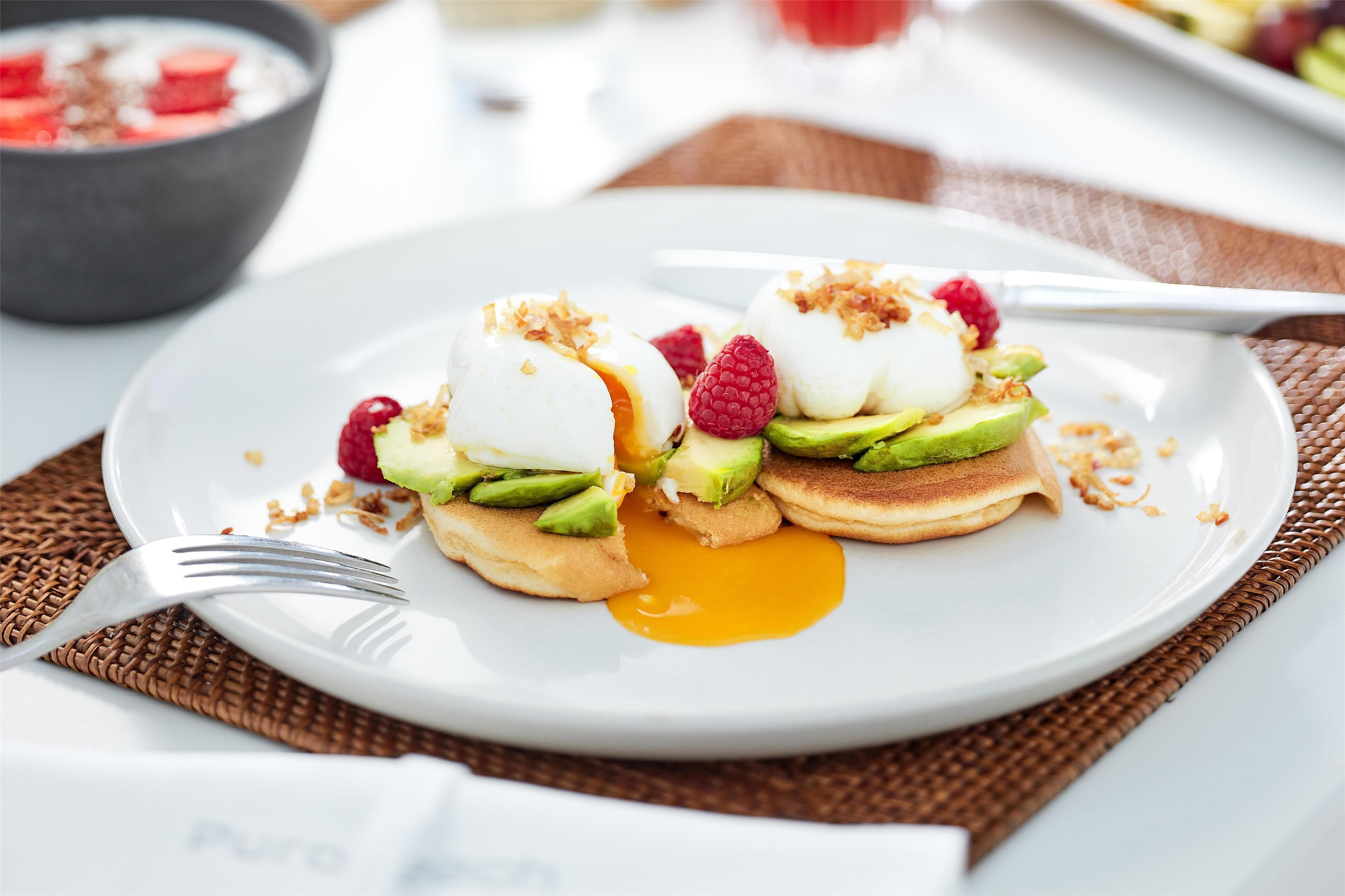 Une assiette de petit-déjeuner avec des crêpes garnies d’avocat, de framboises et d’un œuf poché.