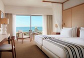 A hotel room with a large bed, a desk with a lamp, a chair, and a view of the ocean through a sliding door.