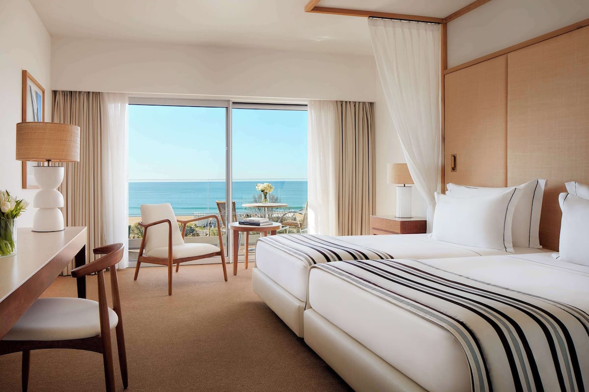 Een hotelkamer met een groot bed, een bureau met lamp, een stoel en uitzicht op de oceaan door een schuifdeur.