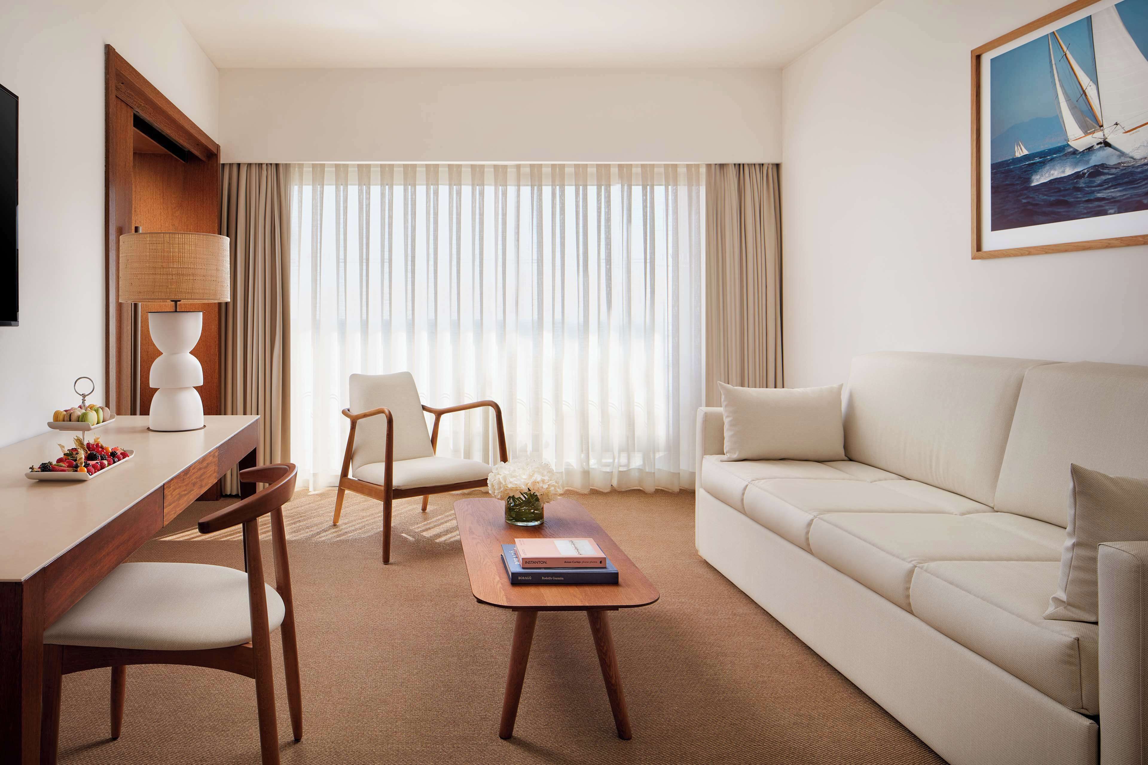 Ein modernes Hotelzimmer mit einem Holztisch, zwei Stühlen, einem Sofa und einem großen Fenster mit transparenten Vorhängen.