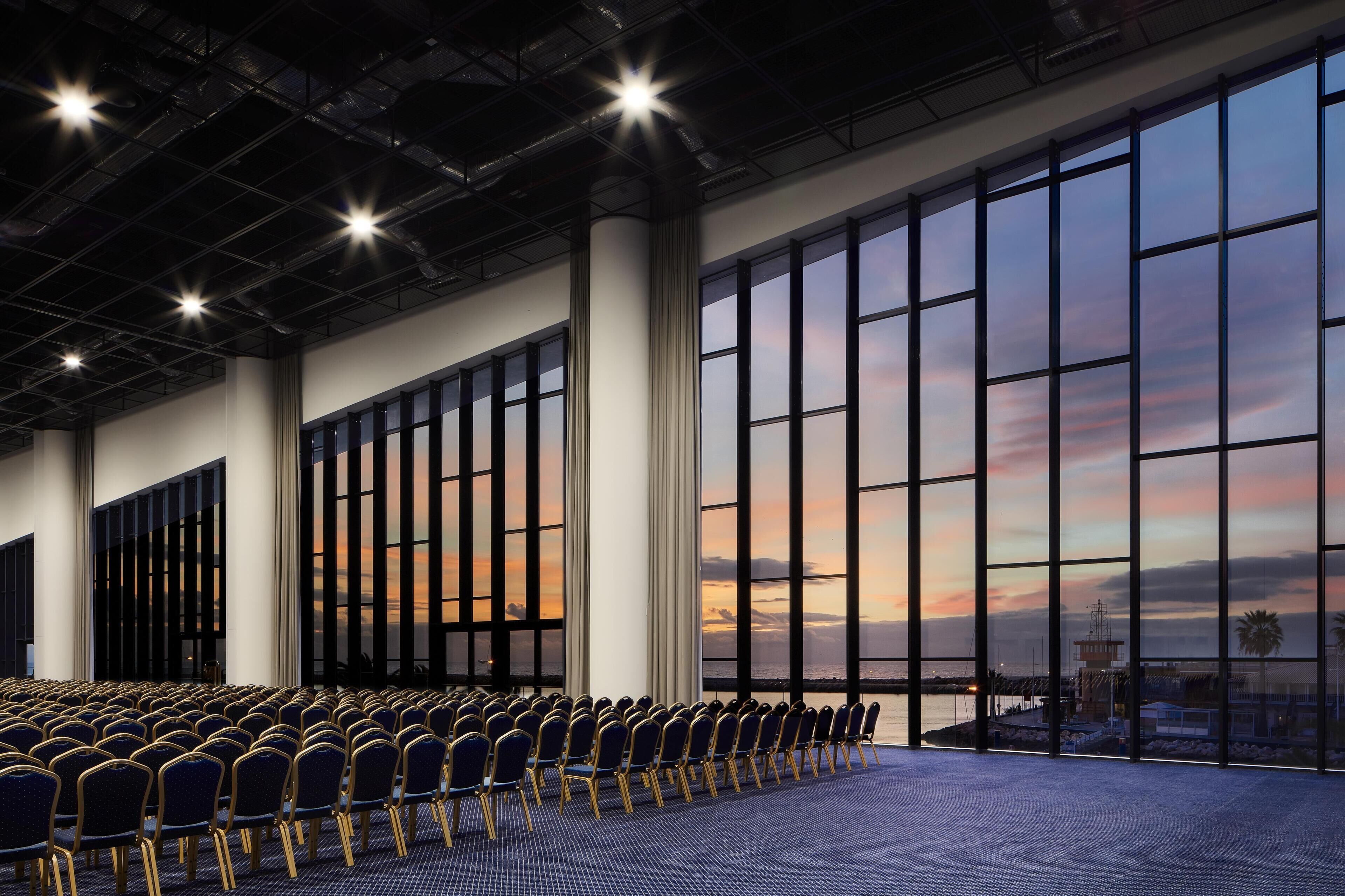A conference room with rows of chairs, large windows, and a view of the sunset.
