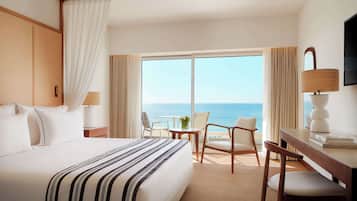 A hotel room with a large bed, a desk with a chair, a TV, and a view of the ocean.