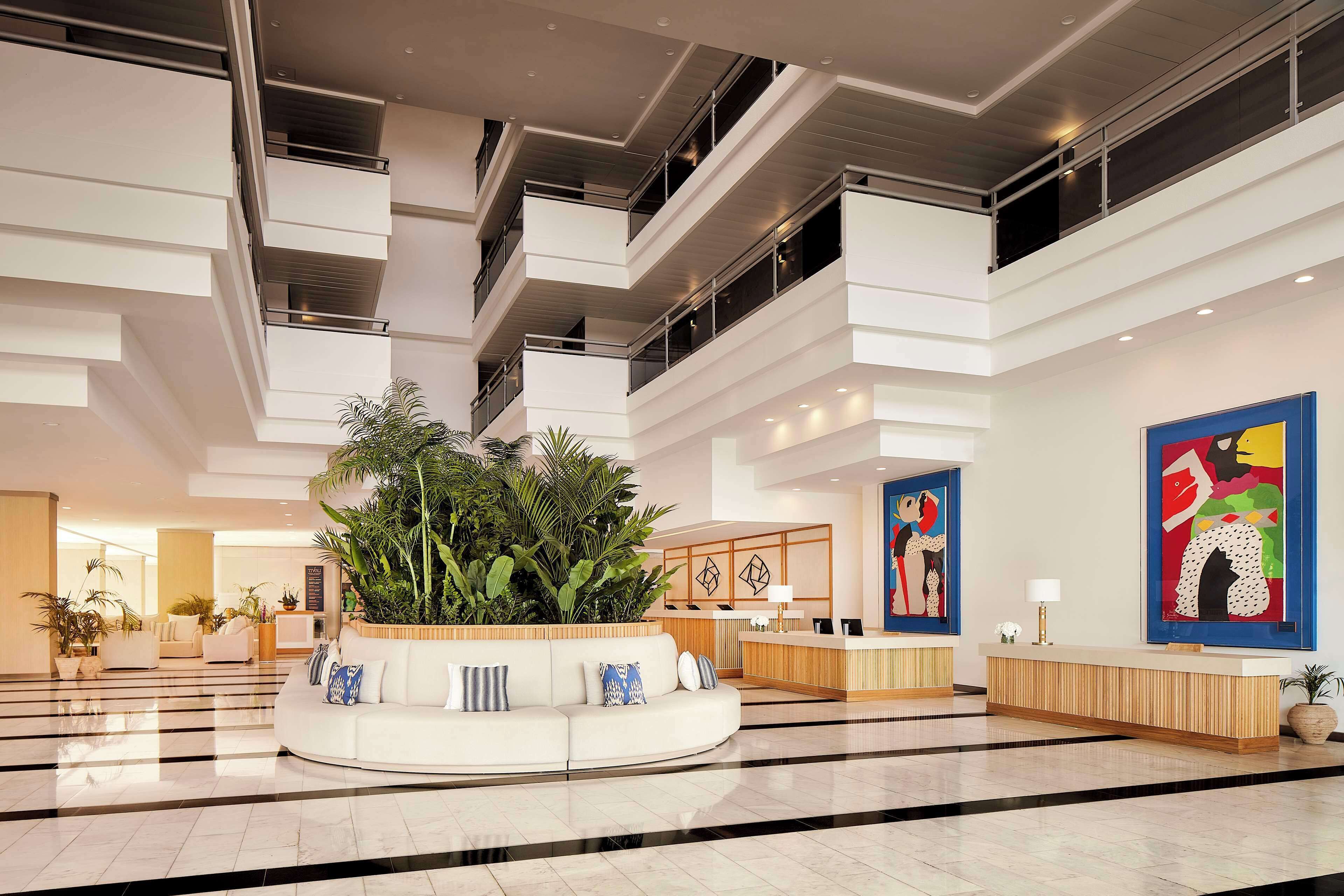 A modern hotel lobby with a central seating area, a reception desk, and a large plant.