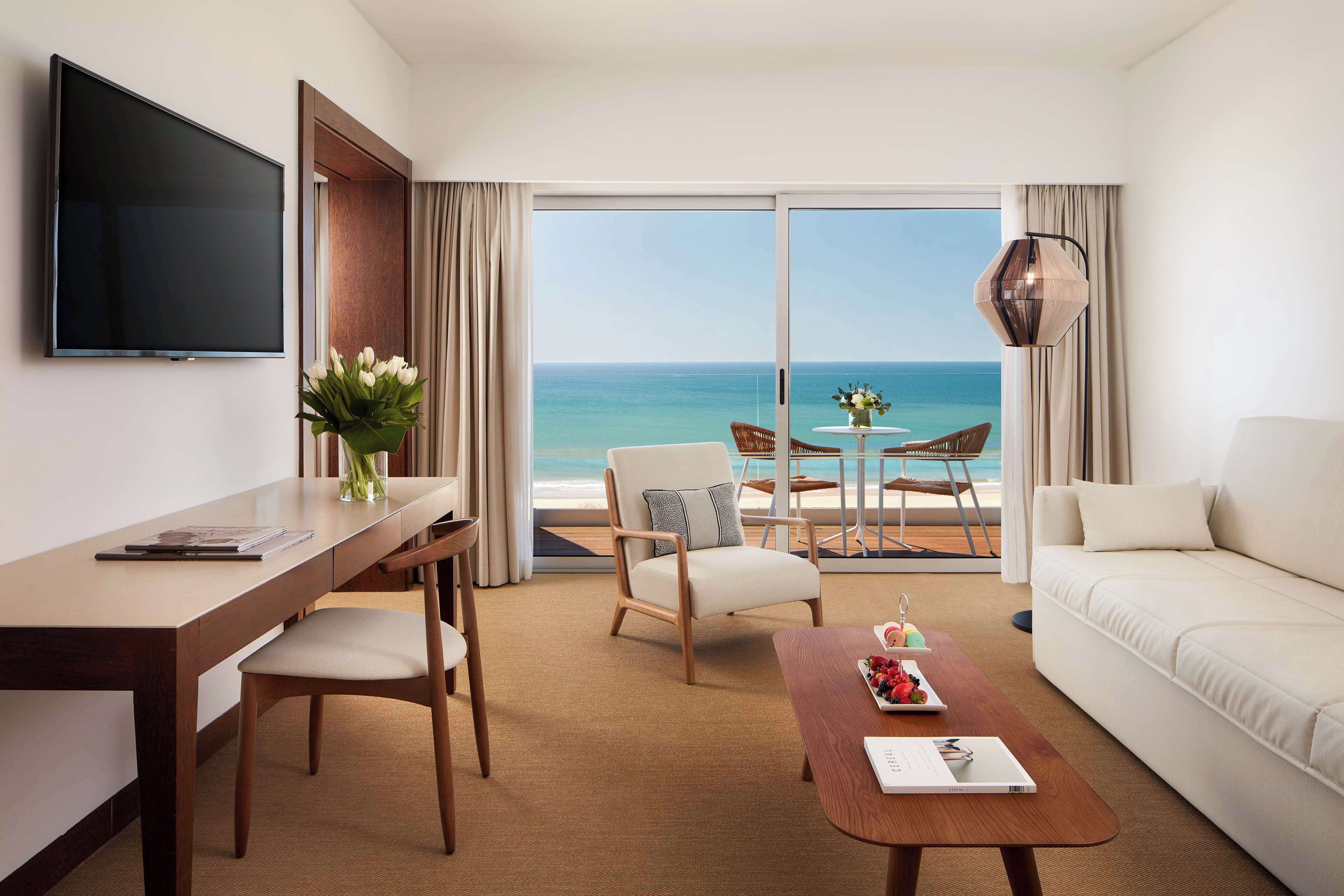 A hotel room with a large TV, a desk with a chair, a sofa, a coffee table with a tray of fruit, and a view of the ocean.