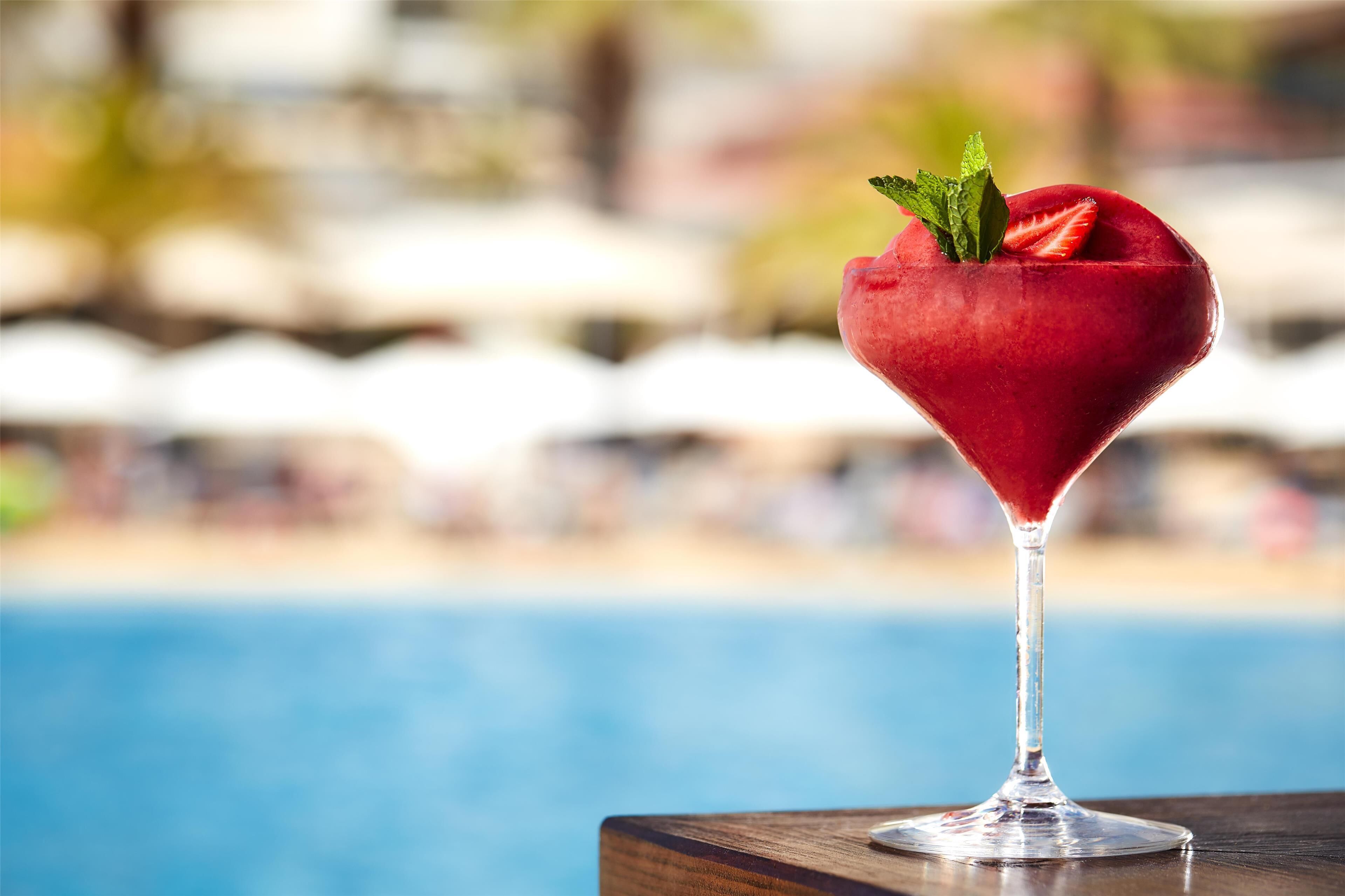 Un cocktail in un bicchiere da martini con decorazione a forma di cuore, guarnito con una fragola e menta, su un tavolo di legno vicino a una piscina.