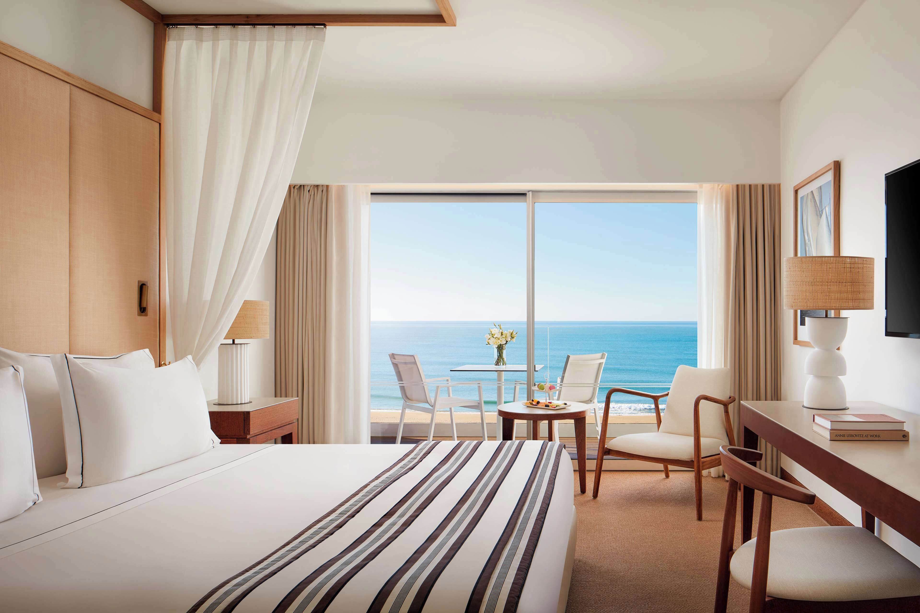 A hotel room with a large bed, a desk, two chairs, a TV, and a view of the ocean.