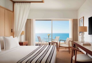 A hotel room with a large bed, a desk, two chairs, a TV, and a view of the ocean.