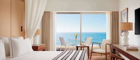 A hotel room with a large bed, a desk, two chairs, a TV, and a view of the ocean.