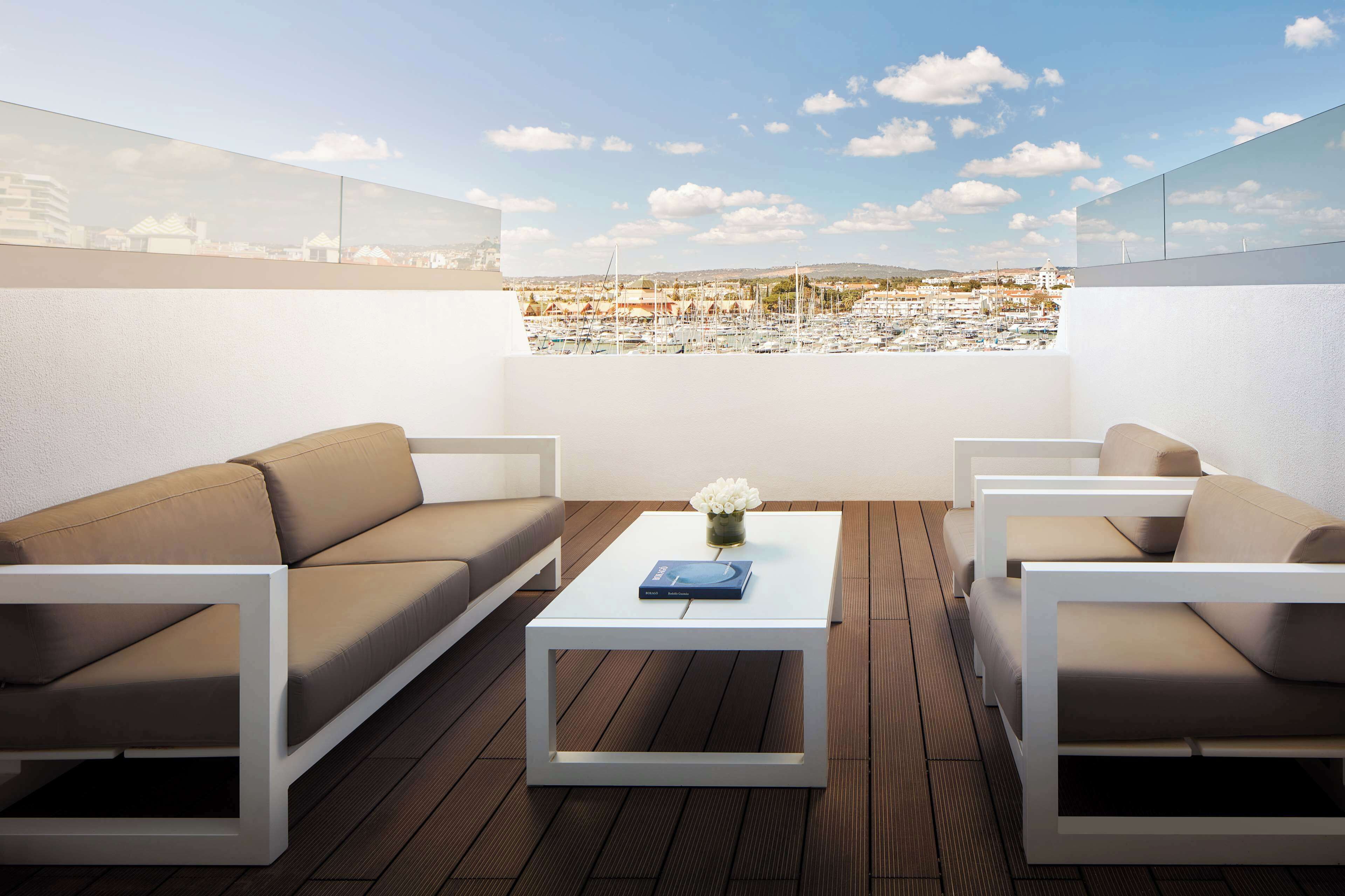 Terraza en la azotea con dos sofás beige, una mesa de centro blanca y vistas a un puerto deportivo y al horizonte de la ciudad.