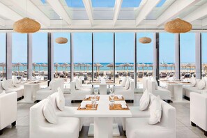 A modern restaurant with white furniture and large windows overlooking a beach.