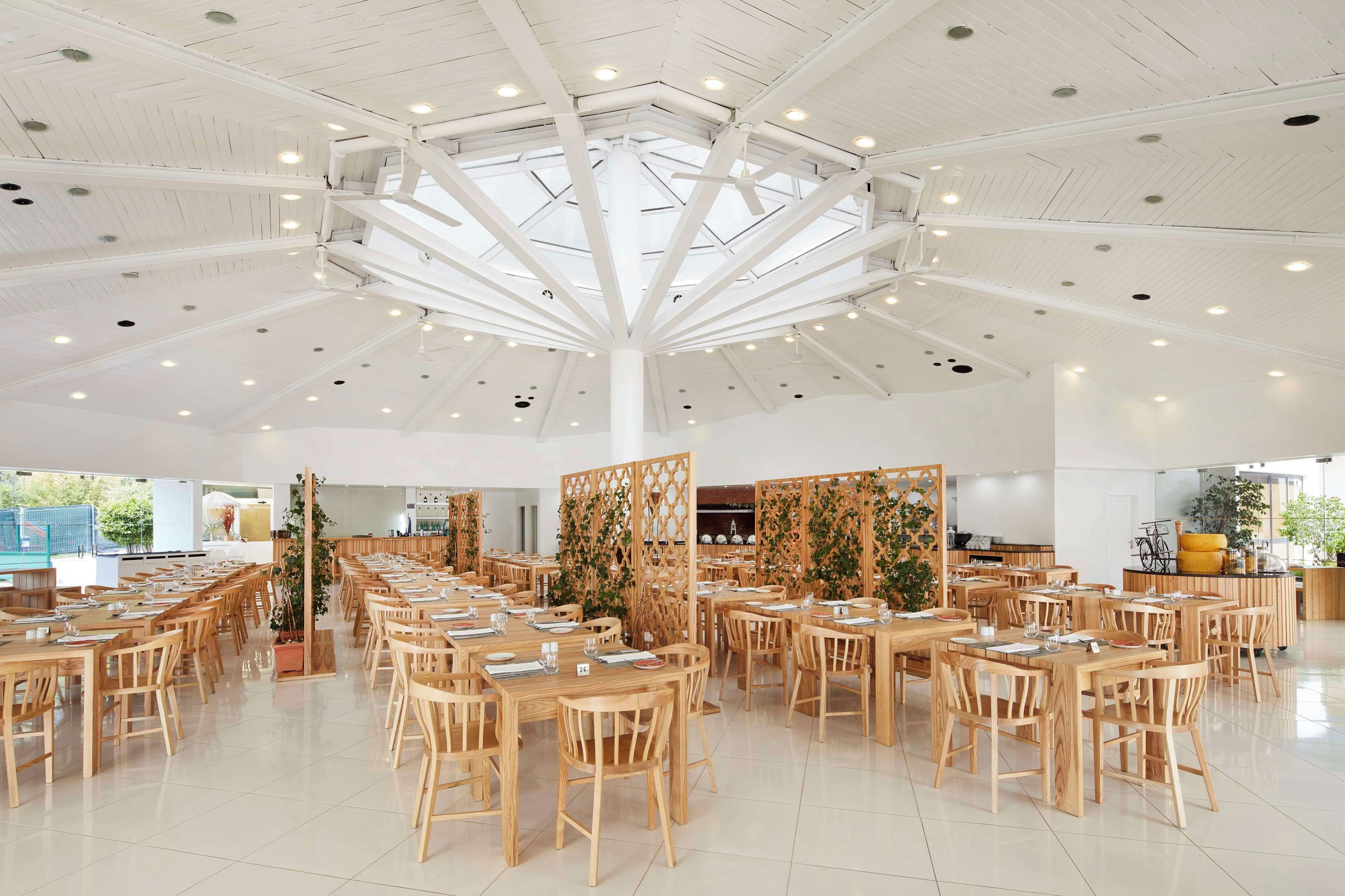 Un comedor amplio con varias mesas y sillas de madera, un techo alto con diseño geométrico y luz natural proveniente de grandes ventanas.