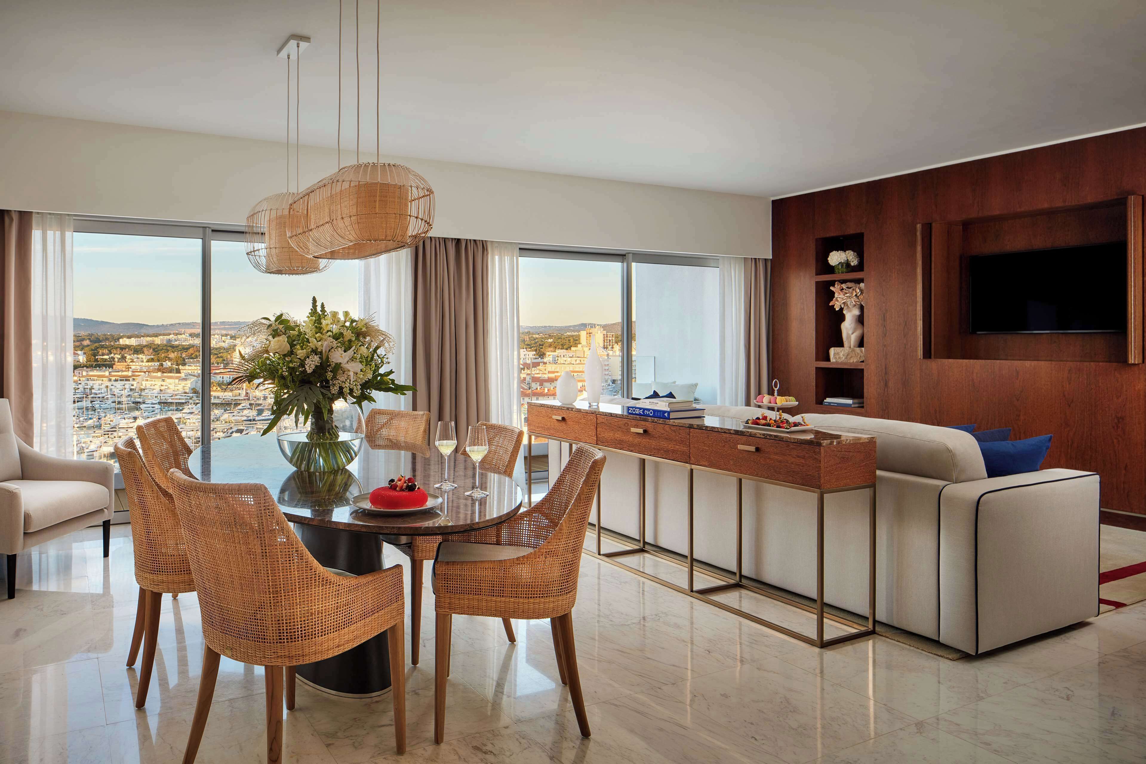 A modern living room with a dining area, a sofa, a TV mounted on a wooden wall, and a large window with a city view.