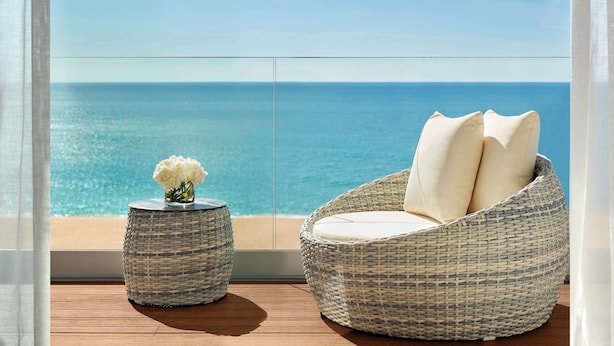 A wicker chair with cushions and a small round table with a vase of flowers on a balcony with a view of the ocean.