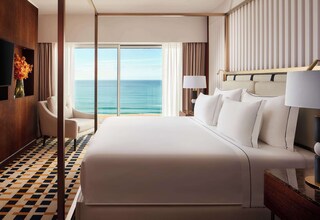 A hotel room with a large bed, a sitting area with a chair, a TV mounted on the wall, and a view of the ocean.