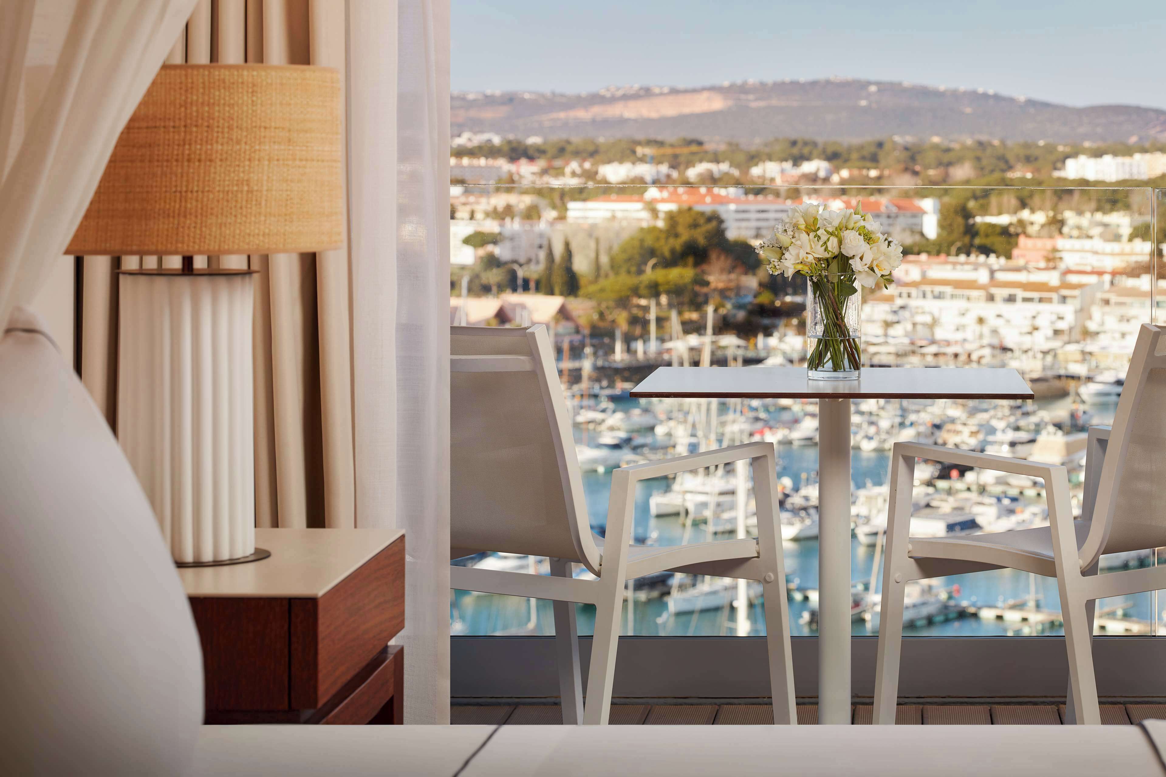A balcony with a table and chairs, a vase of flowers, and a view of a marina and buildings.