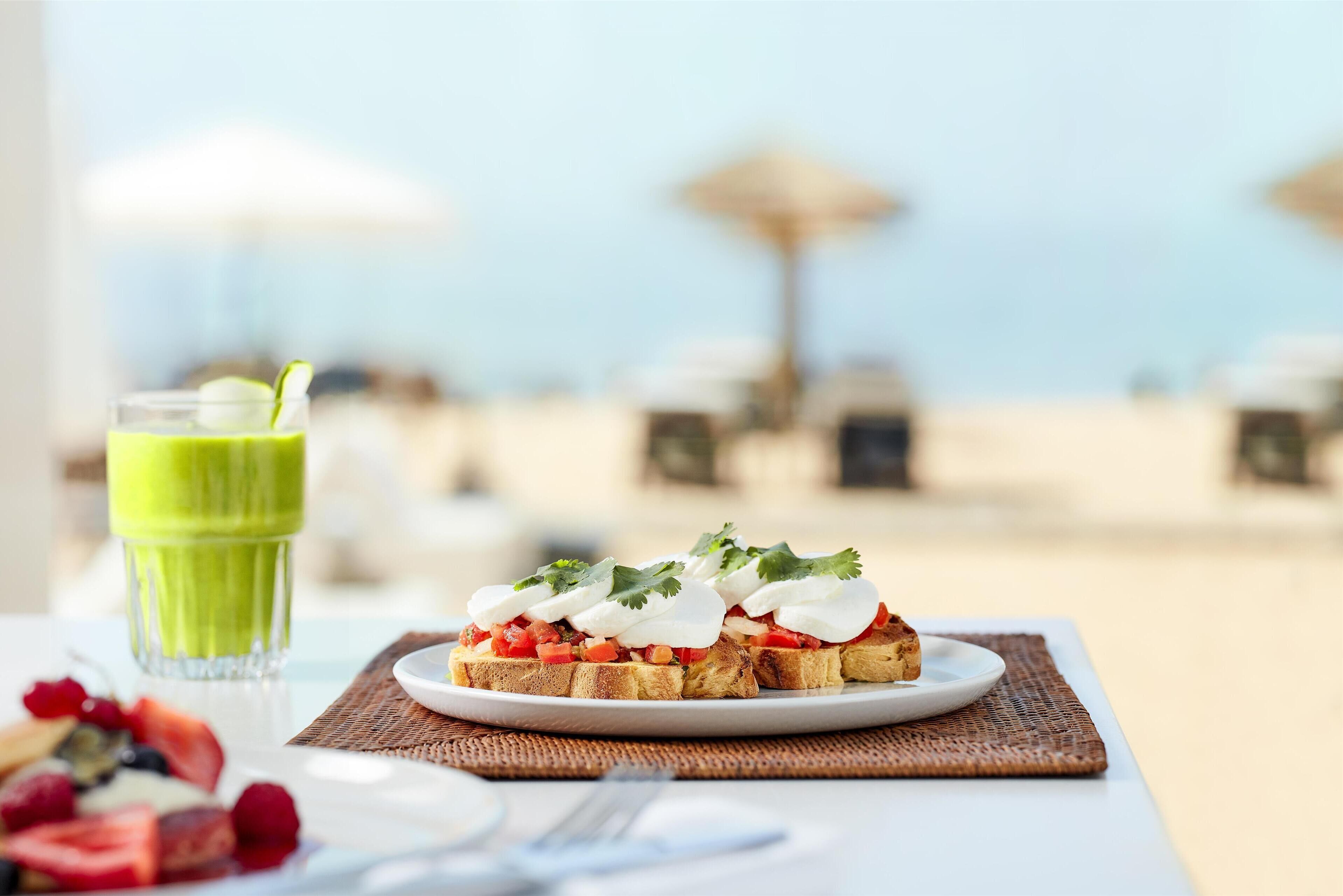 A plate of toast with tomatoes, cream, and cilantro, a glass of green smoothie, and a wicker placemat on a table with a view of a beach and umbrellas in the background.