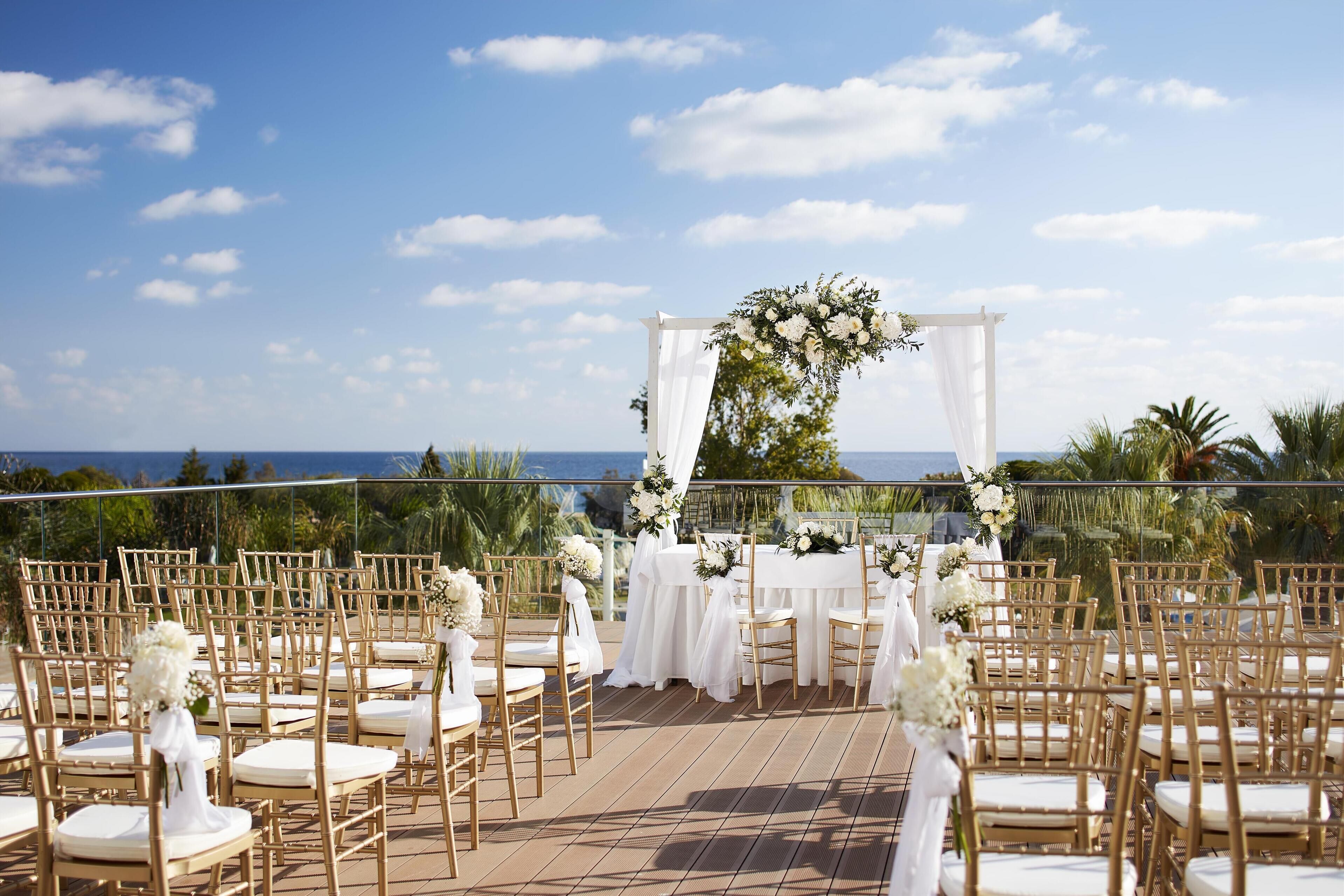 An outdoor wedding ceremony setup with white floral arrangements, gold chairs, and a view of the ocean.
