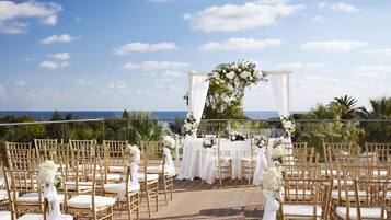 An outdoor wedding ceremony setup with white floral arrangements, gold chairs, and a view of the ocean.