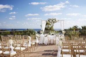 An outdoor wedding ceremony setup with white floral arrangements, gold chairs, and a view of the ocean.