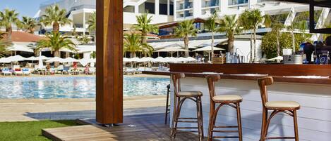 A wooden deck with three bar stools, a pool, and a view of a resort with palm trees and umbrellas.