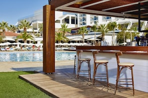 A wooden deck with three bar stools, a pool, and a view of a resort with palm trees and umbrellas.