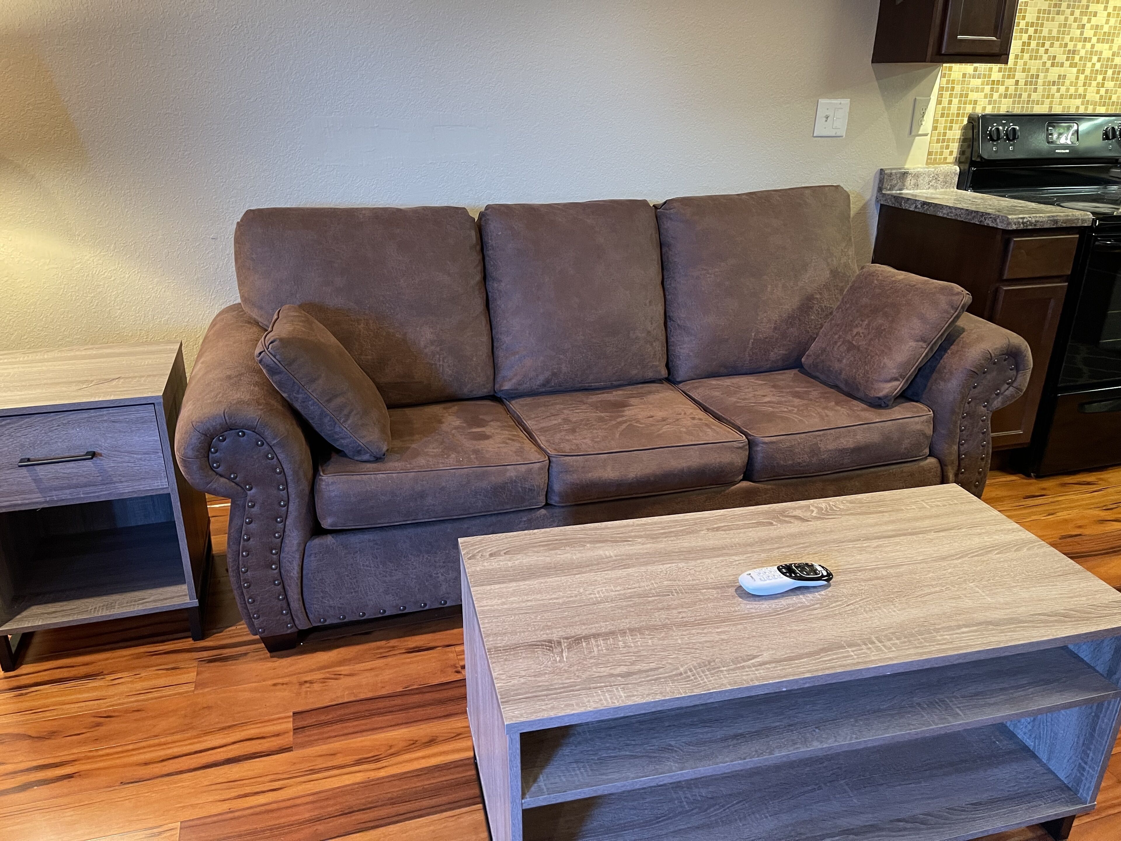 Una sala de estar con un sofá marrón, una mesa de centro de madera y una cocina al fondo.