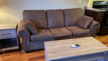 Una sala de estar con un sofá marrón, una mesa de centro de madera y una cocina al fondo.