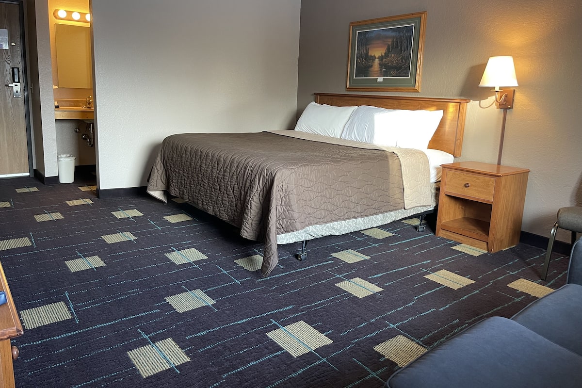 Ein Hotelzimmer mit Bett, Nachttisch, Lampe, einem Gemälde an der Wand und einem Badezimmer mit Waschbecken und Spiegel.