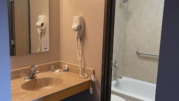 A bathroom with a wooden vanity, a sink, a mirror with lights, a hairdryer, and a shower area.