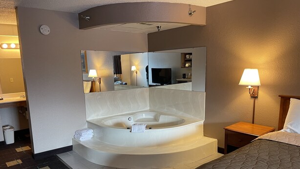 A hotel room with a large bathtub, a bed, a nightstand, a lamp, a mirror, and a bathroom with a sink and toilet. - Walker Hotel (Walker)