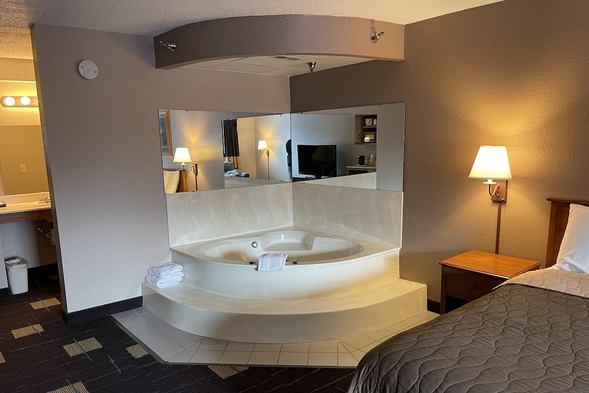 Ein Hotelzimmer mit einer großen Badewanne, einem Bett, einem Nachttisch, einer Lampe, einem Spiegel und einem Badezimmer mit Waschbecken und Toilette.