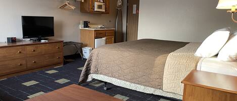 A hotel room with a bed, a TV, a wooden dresser, a desk with a microwave, and a lamp.