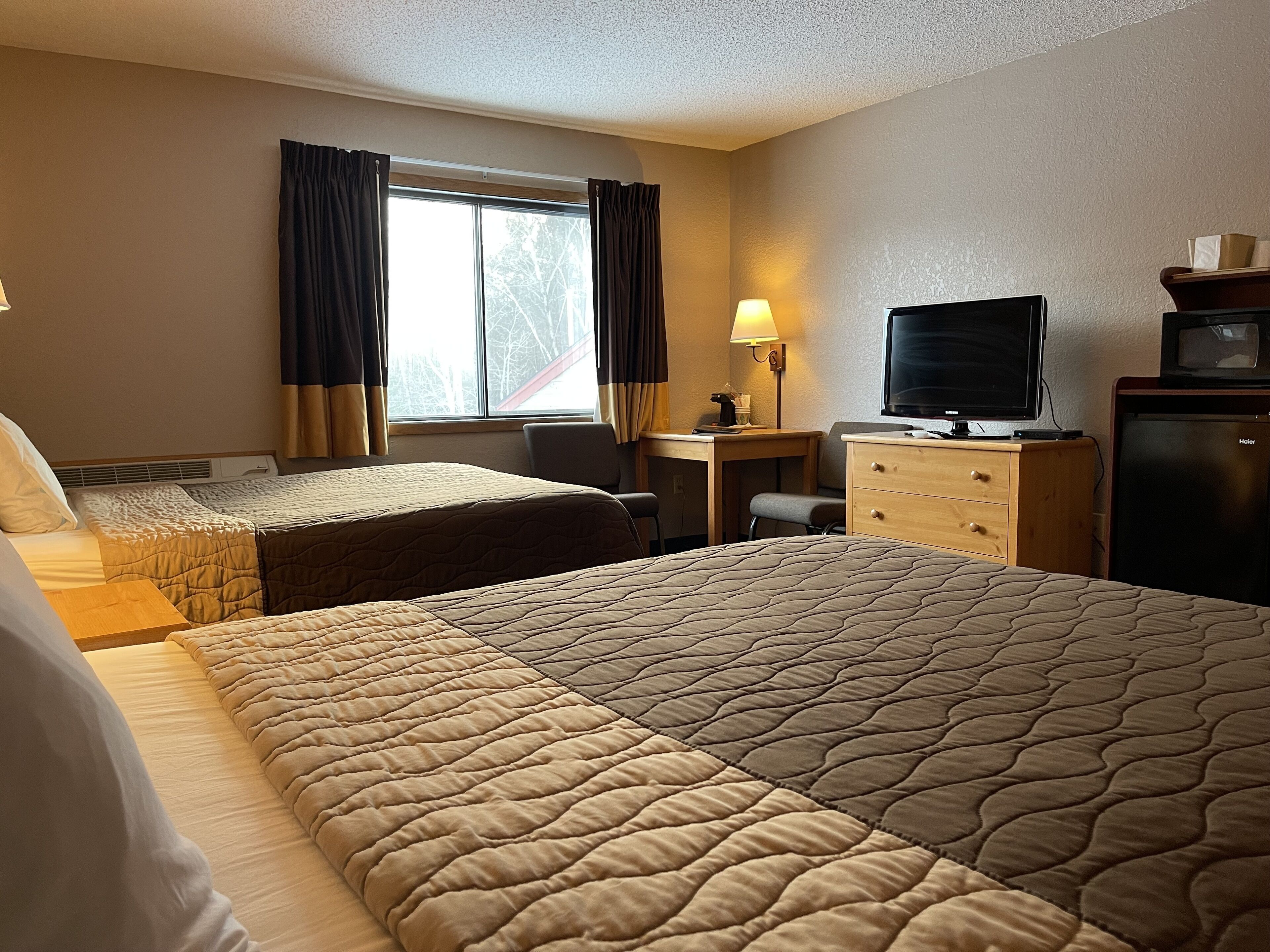 A hotel room with two beds, a desk with a lamp, a television, and a refrigerator.