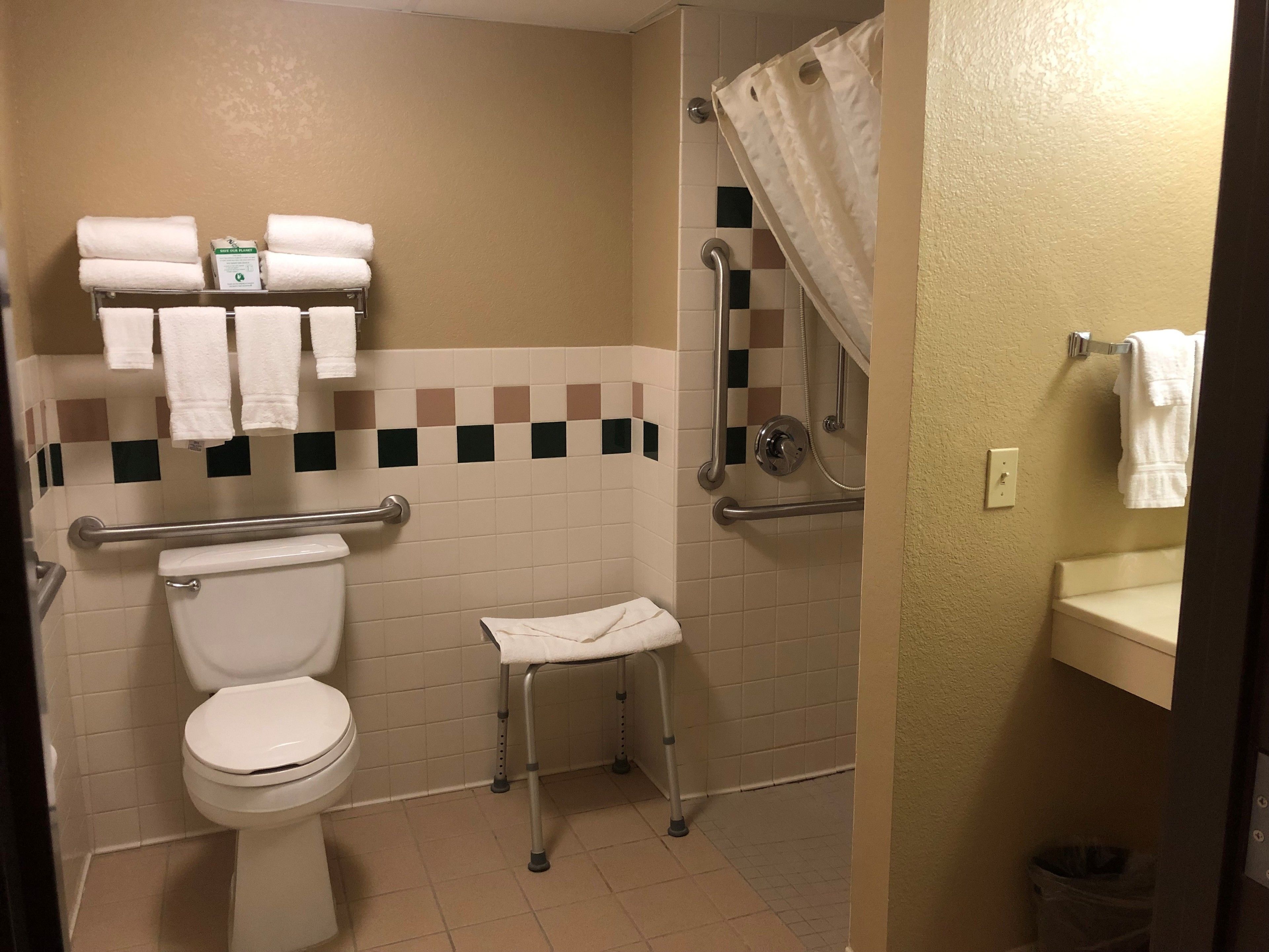 A bathroom with a toilet, a shower with a grab bar, a small stool, and a towel rack with towels.