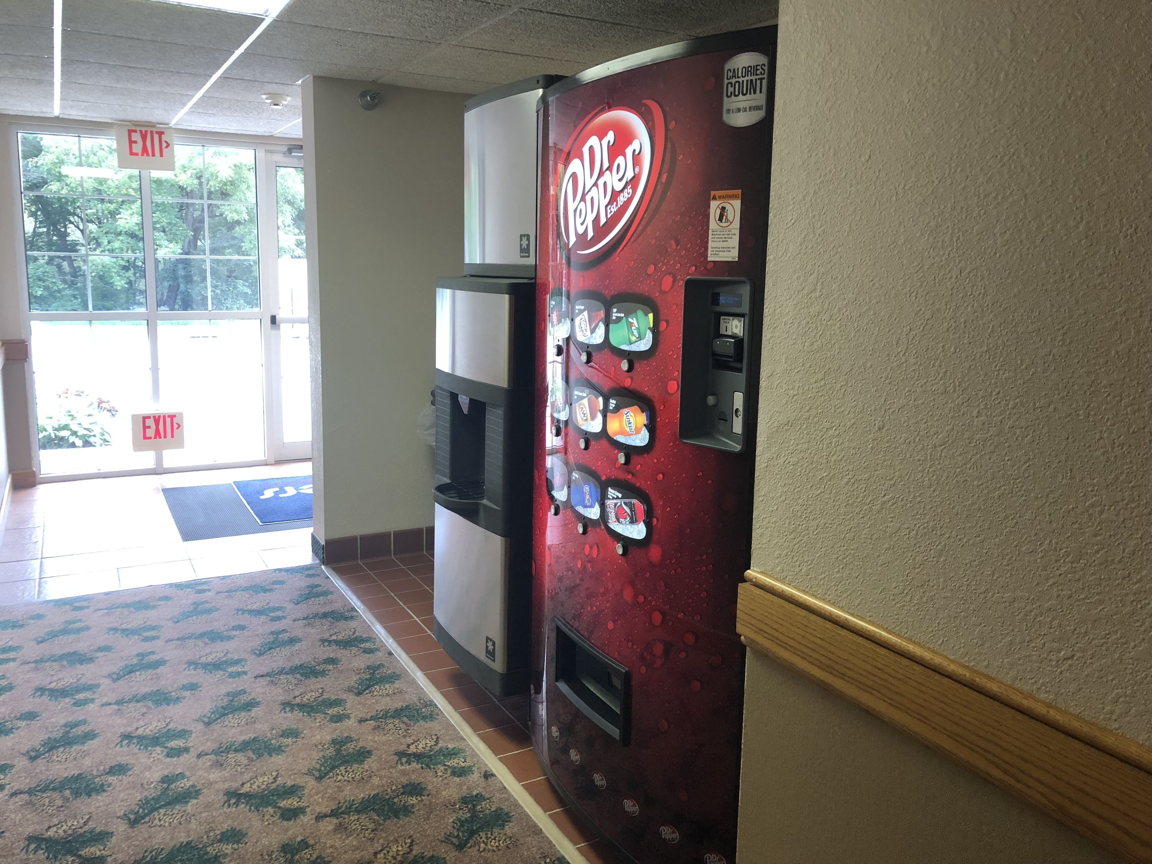 A Dr Pepper vending machine in a hallway with an exit sign and a patterned carpet.