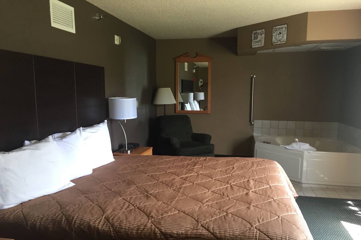 A hotel room with a large bed, a chair, a desk, a lamp, a mirror, and a bathtub.