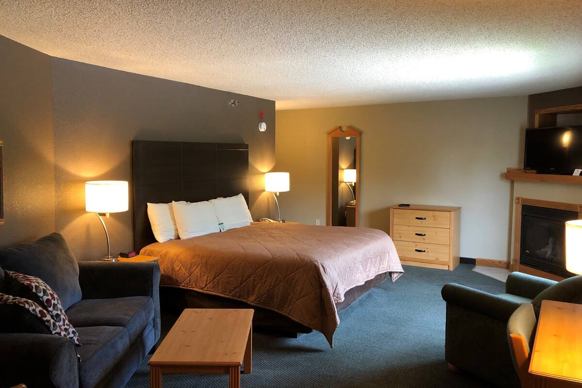 A hotel room with a bed, a sofa, a small table, a dresser, a TV, and a fireplace.