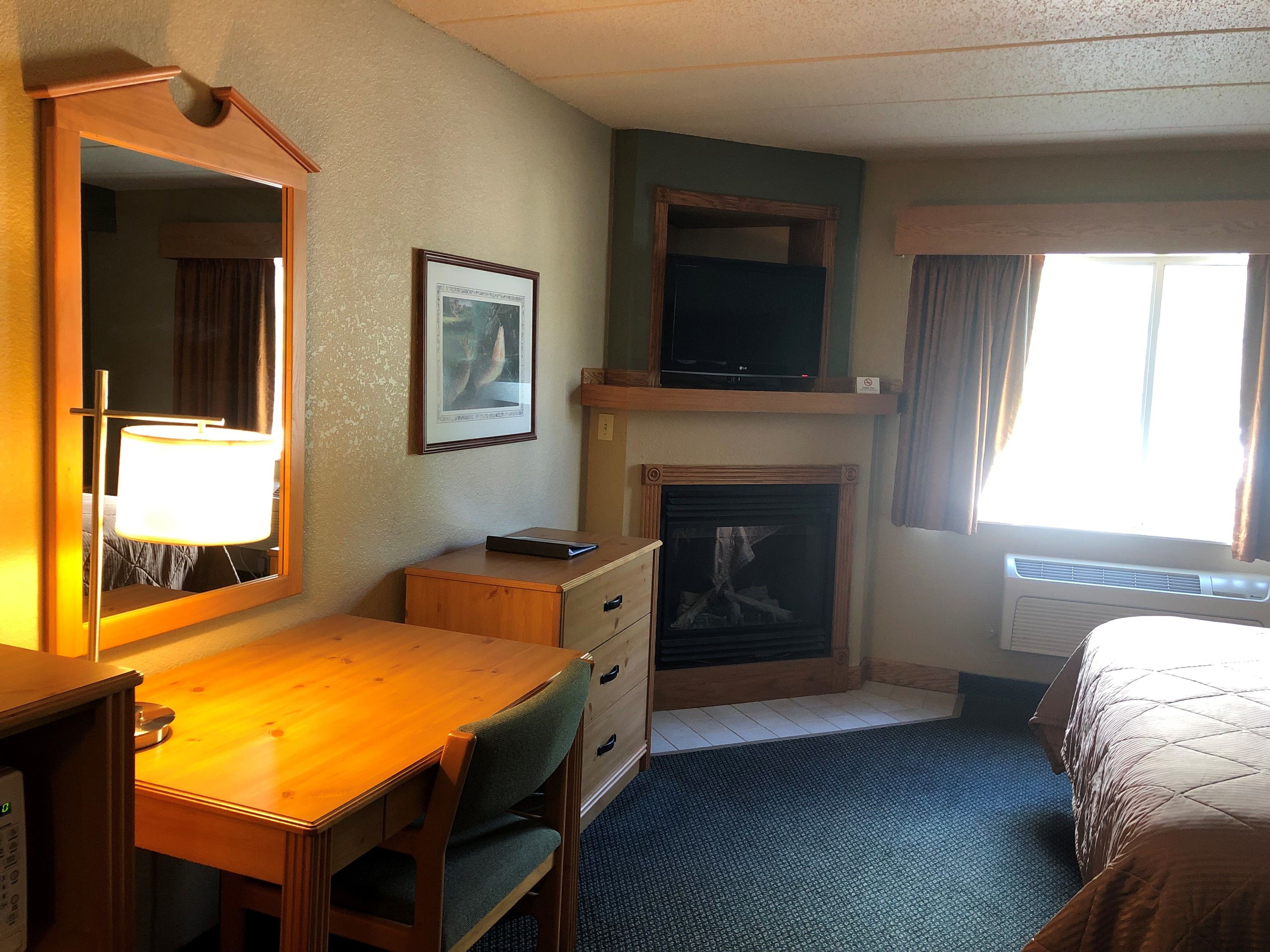 A hotel room with a wooden desk, a chair, a dresser, a television, a fireplace, and a bed.