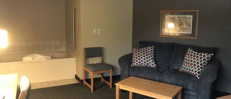 A hotel room with a bathtub, a wooden chair, a dark-colored sofa with patterned cushions, and a small wooden table.