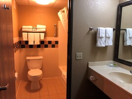 Baño con inodoro, lavamanos y espejo. Hay toallas colgadas en un perchero y una cortina de ducha.
