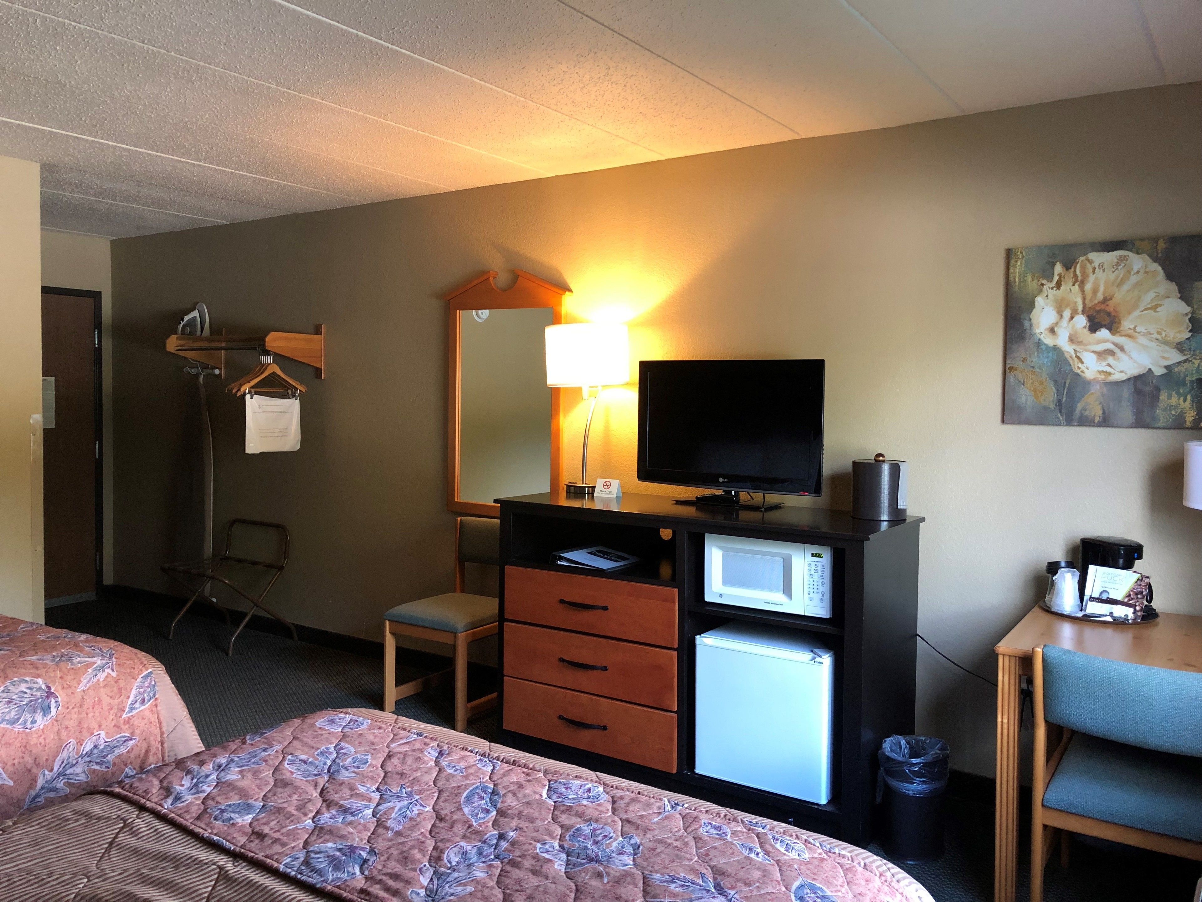 Habitación de hotel con cama, escritorio con computadora, refrigerador pequeño, televisor, lámpara y un cuadro en la pared.