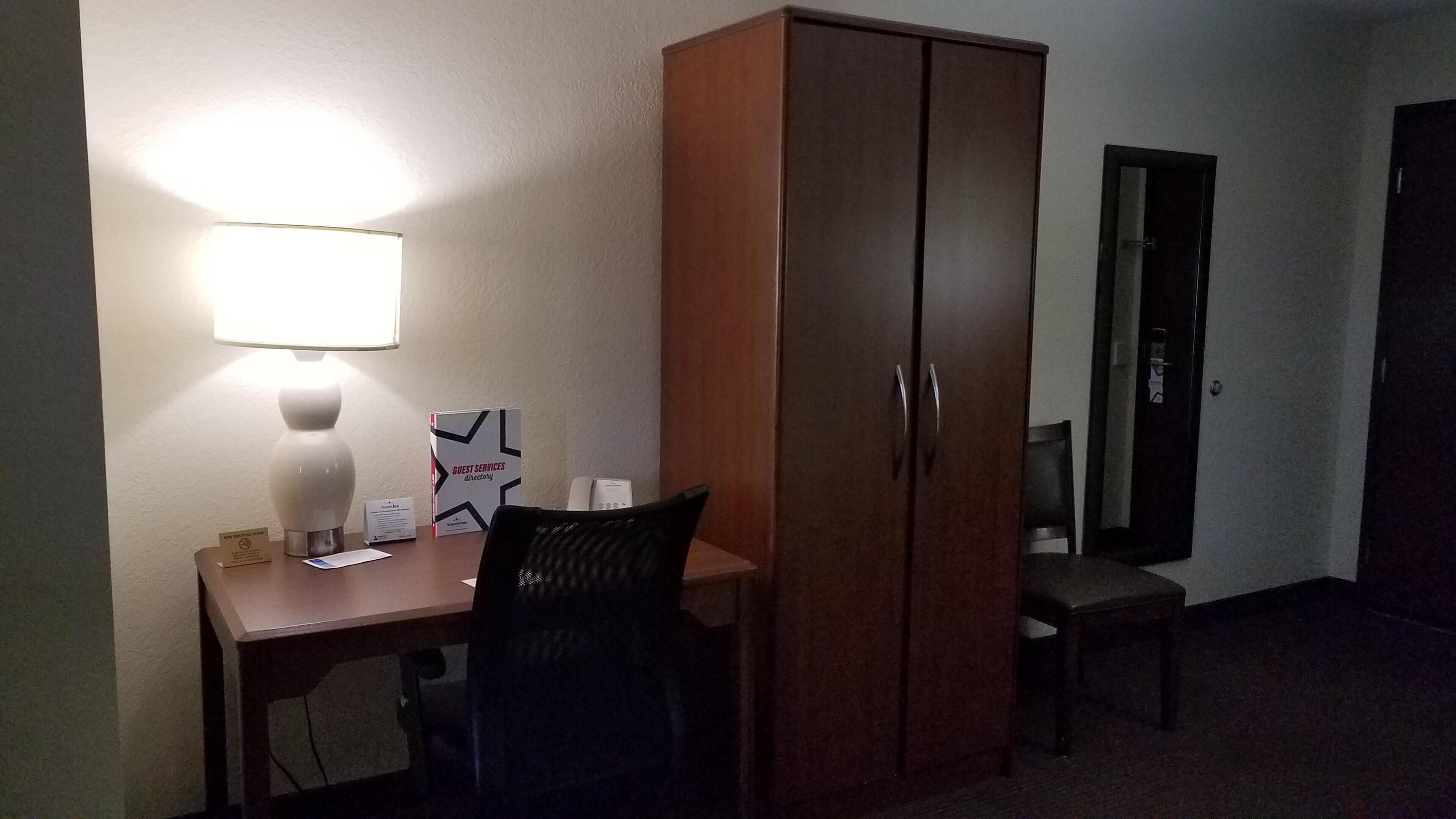 A hotel room with a desk, a chair, a lamp, a wardrobe, and a door.