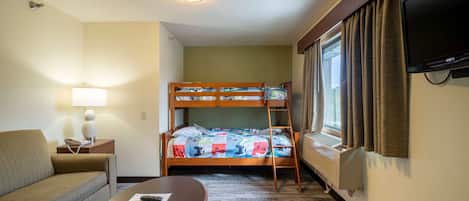 A hotel room with a bunk bed, a sofa, a table, a lamp, a TV, and a window with curtains.