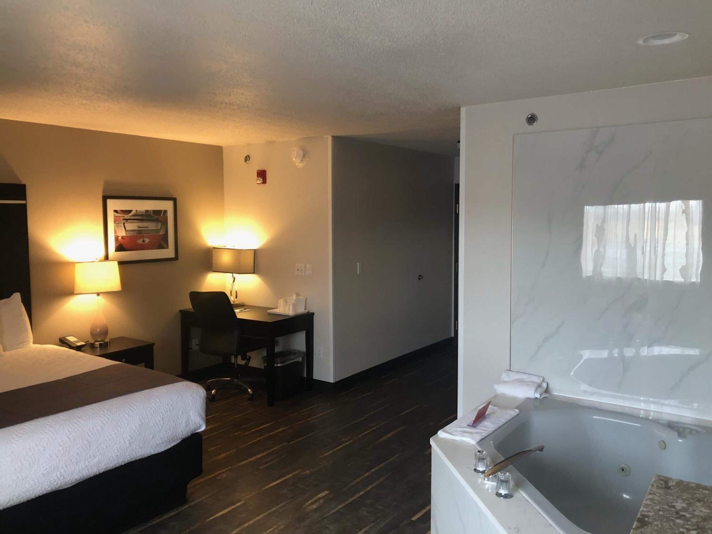 A hotel room with a bed, desk, chair, two lamps, and a bathtub.