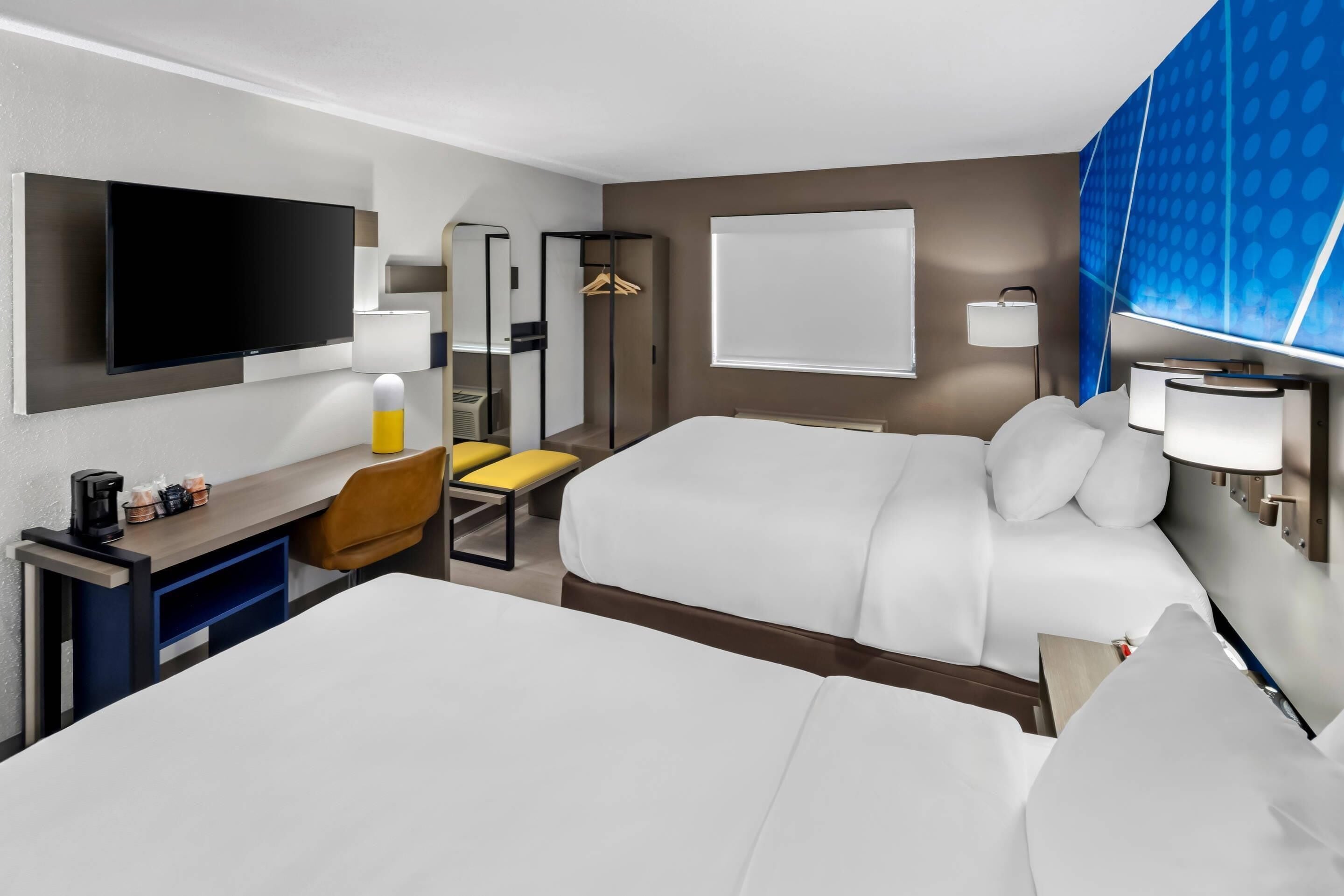 A hotel room with a bed, a desk with a chair, a TV, a mirror, a wardrobe, and a window.