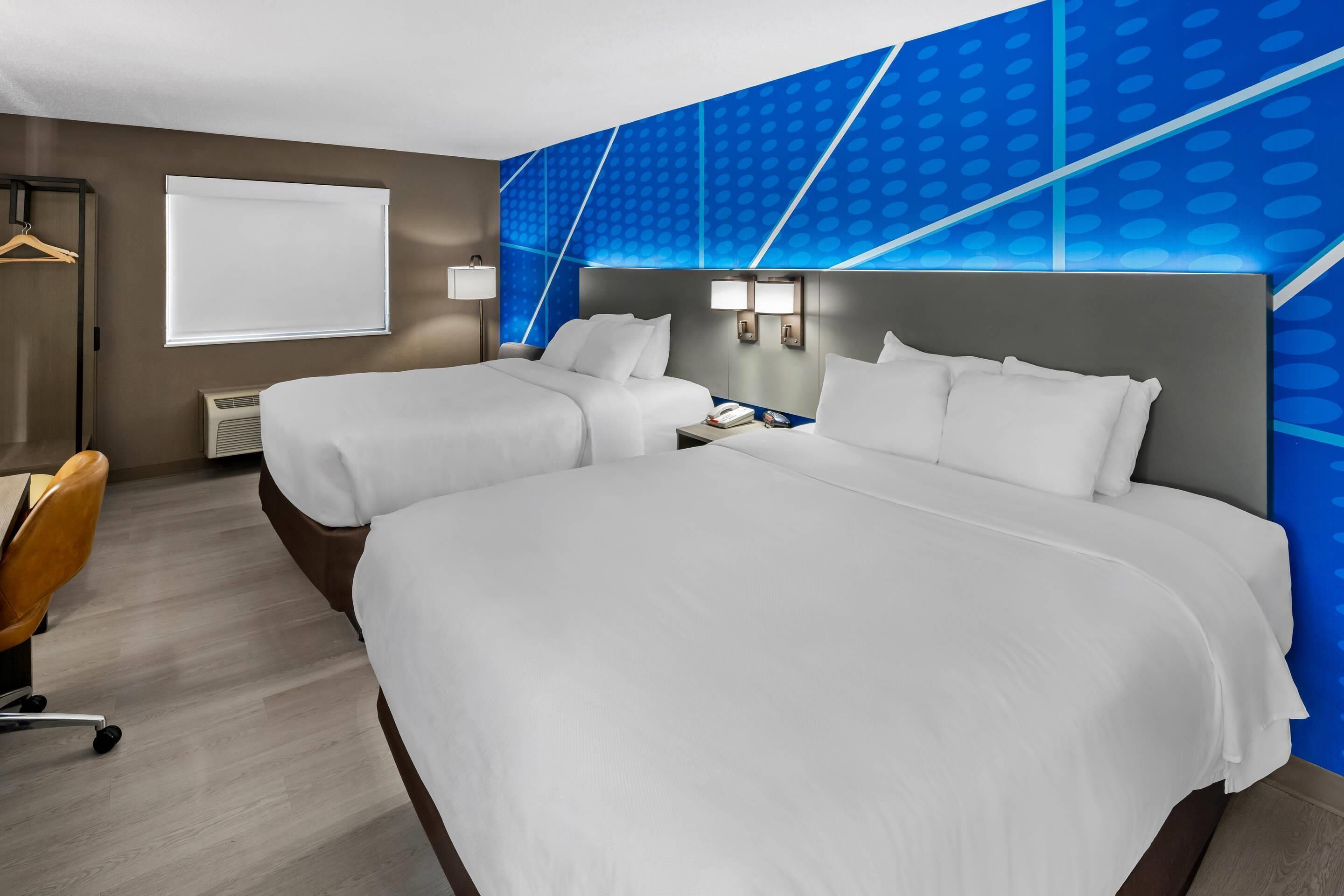 A hotel room with two beds, a desk, a chair, a window, and a wall with a geometric pattern.