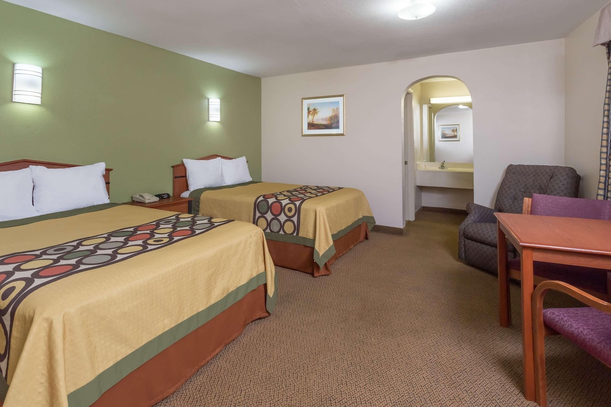 A hotel room with two beds, a desk, a chair, and a bathroom visible through an archway.