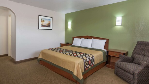 Una habitación de hotel con cama, una silla, una mesita de noche, una lámpara y un cuadro en la pared.