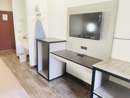A modern hotel room with a flat-screen TV mounted on the wall, a small desk with a chair, and a trash bin.